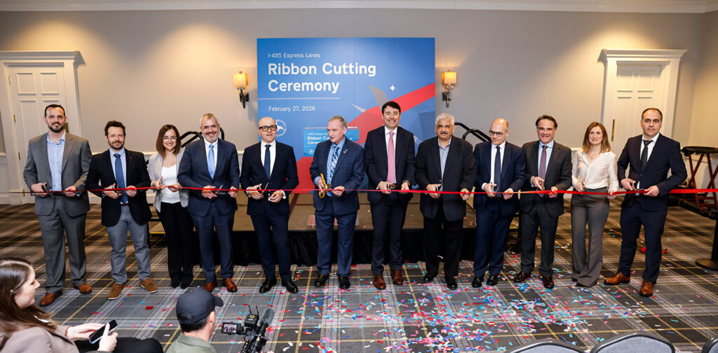 A group of 12 well-dressed professionals hold a red ribbon, smiling, at the I-485 Express Lane opening. The man in the left middle poses with gold scissors.